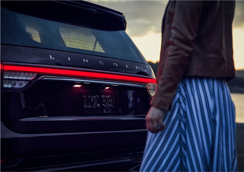 A person is shown near the rear of a 2023 Lincoln Aviator® SUV as the Lincoln Embrace illuminates the rear lights | Dan Cummins Lincoln in Nicholasville KY