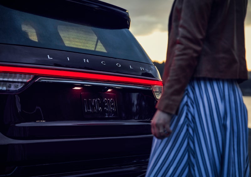 A person is shown near the rear of a 2024 Lincoln Aviator® SUV as the Lincoln Embrace illuminates the rear lights | Dan Cummins Lincoln in Nicholasville KY
