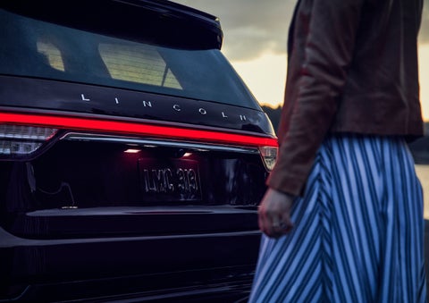 A person is shown near the rear of a 2024 Lincoln Aviator® SUV as the Lincoln Embrace illuminates the rear lights | Dan Cummins Lincoln in Nicholasville KY